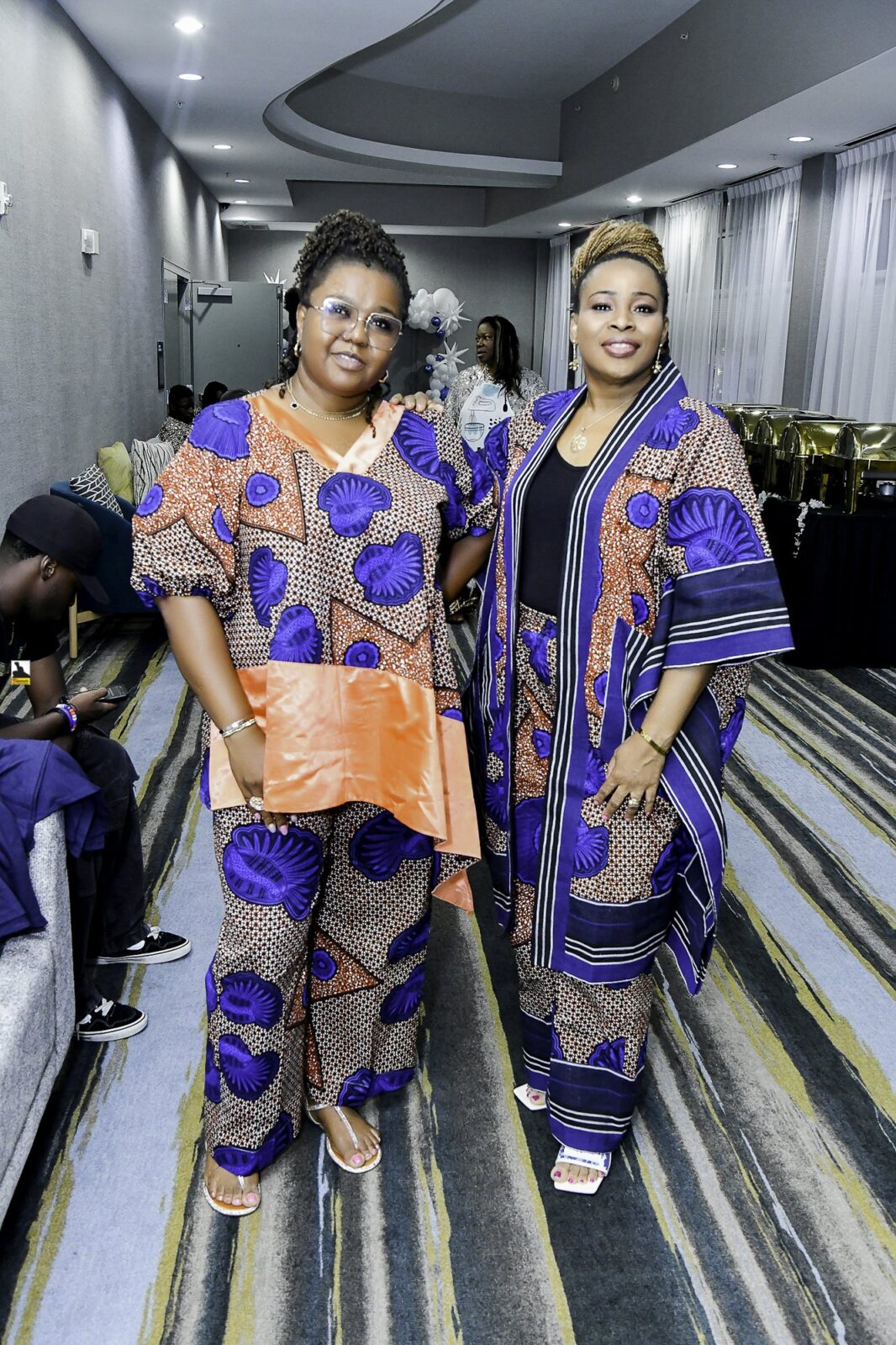 Two women in patterned clothing seated together, one wearing orange