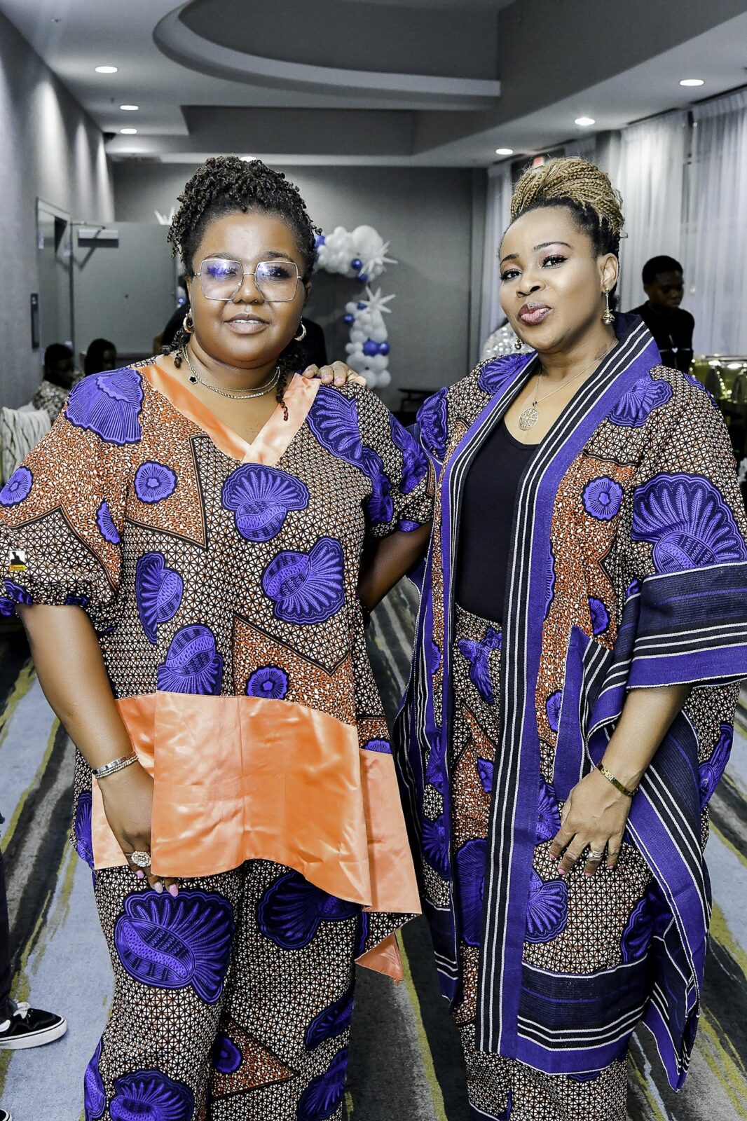 Two women in patterned clothing seated together, one wearing orange
