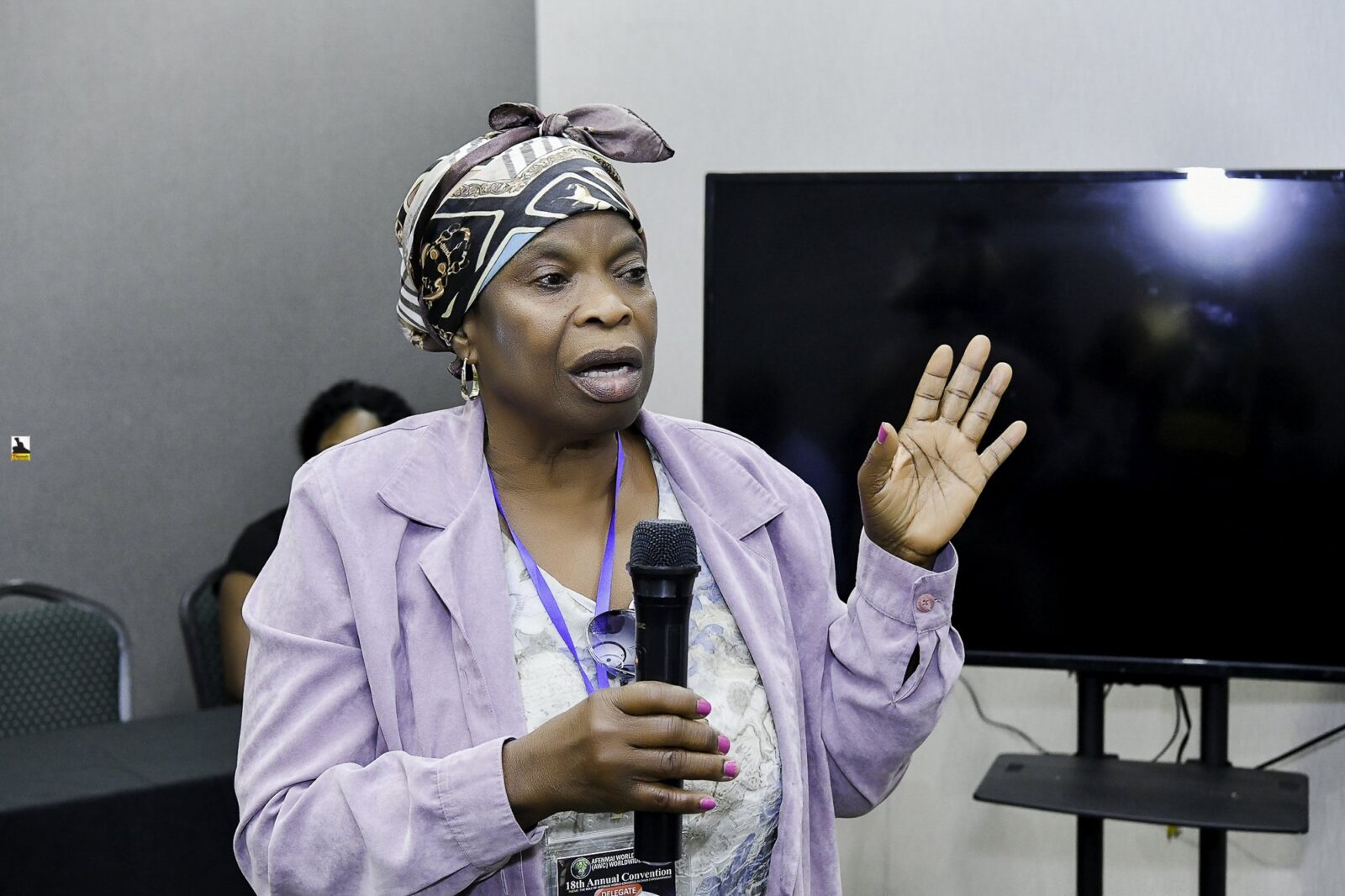 Woman in a lavender jacket holding a microphone and raising her hand