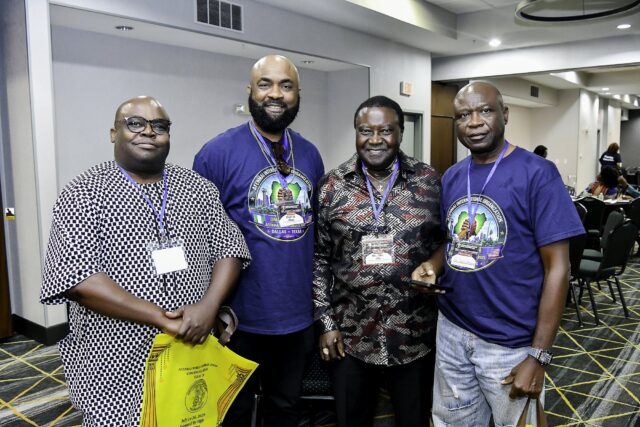 Four men in blue shirts and patterned clothing smiling together