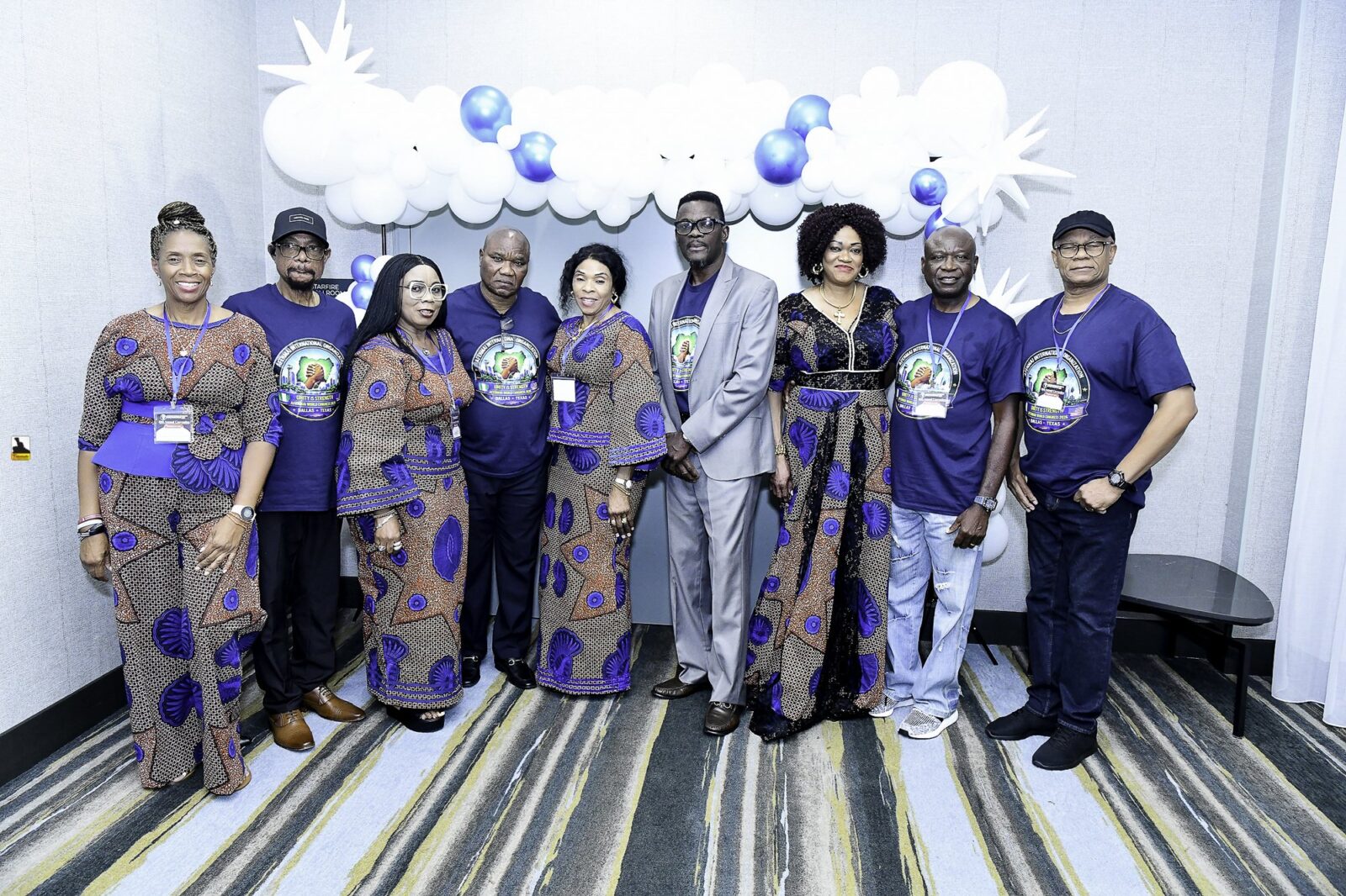 Nine attendees in blue shirts and patterned outfits beneath a balloon arch