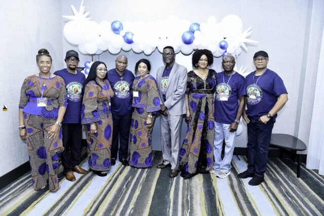 Nine attendees in blue shirts and patterned outfits beneath a balloon arch