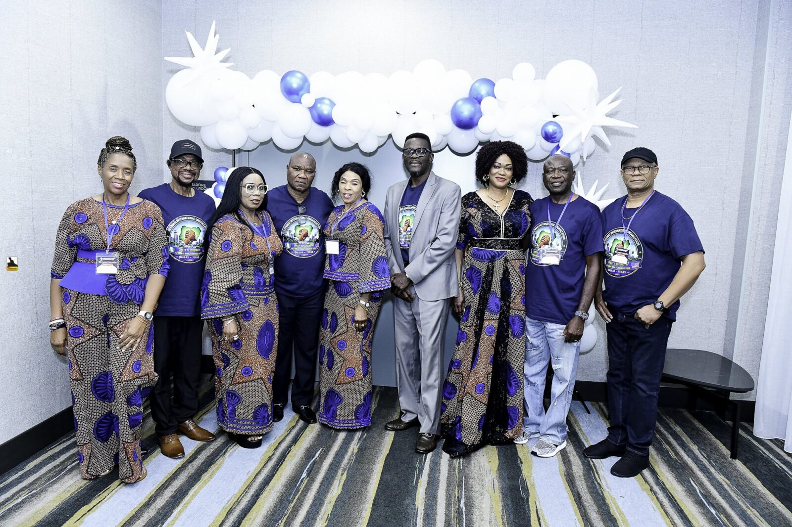 Nine attendees in blue shirts and patterned outfits beneath a balloon arch