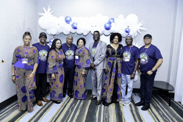 Nine attendees in blue shirts and patterned outfits beneath a balloon arch
