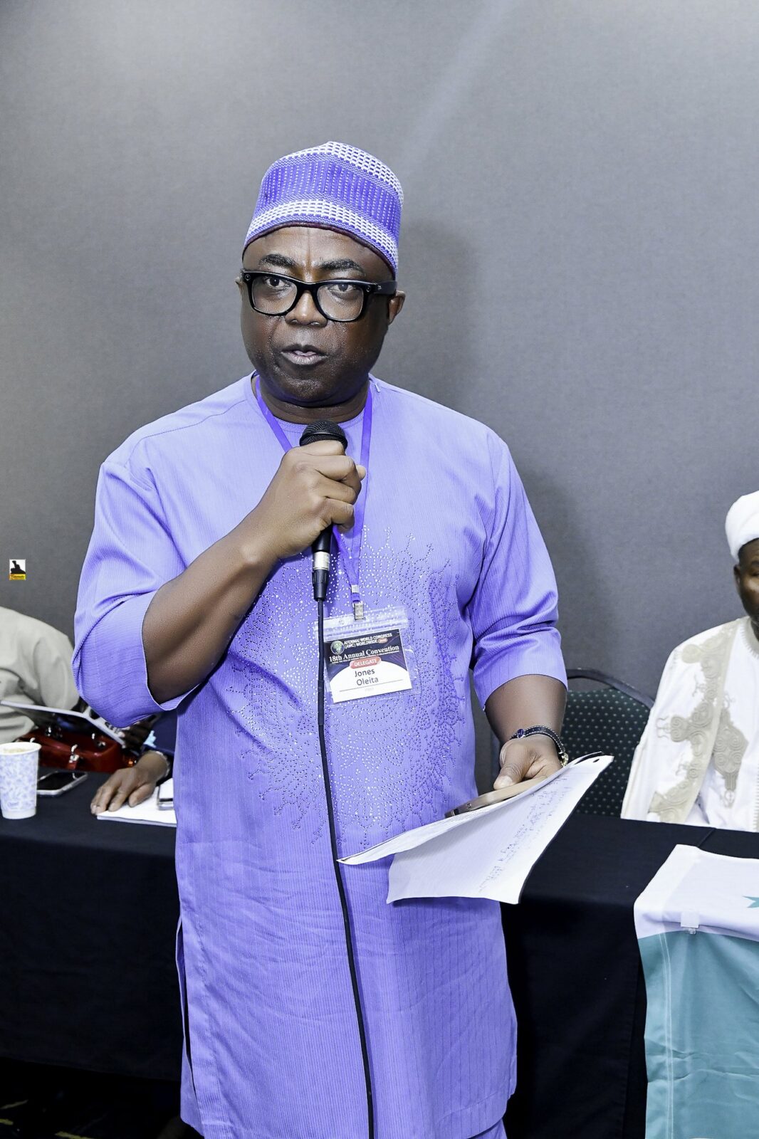 Man in lavender clothing and a cap speaking into a microphone