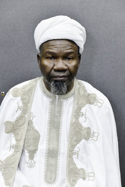 Man in embroidered white clothing and a white cap seated in the room