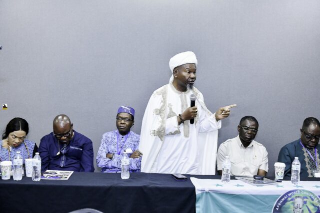 Man in embroidered white clothing addressing attendees at the front table