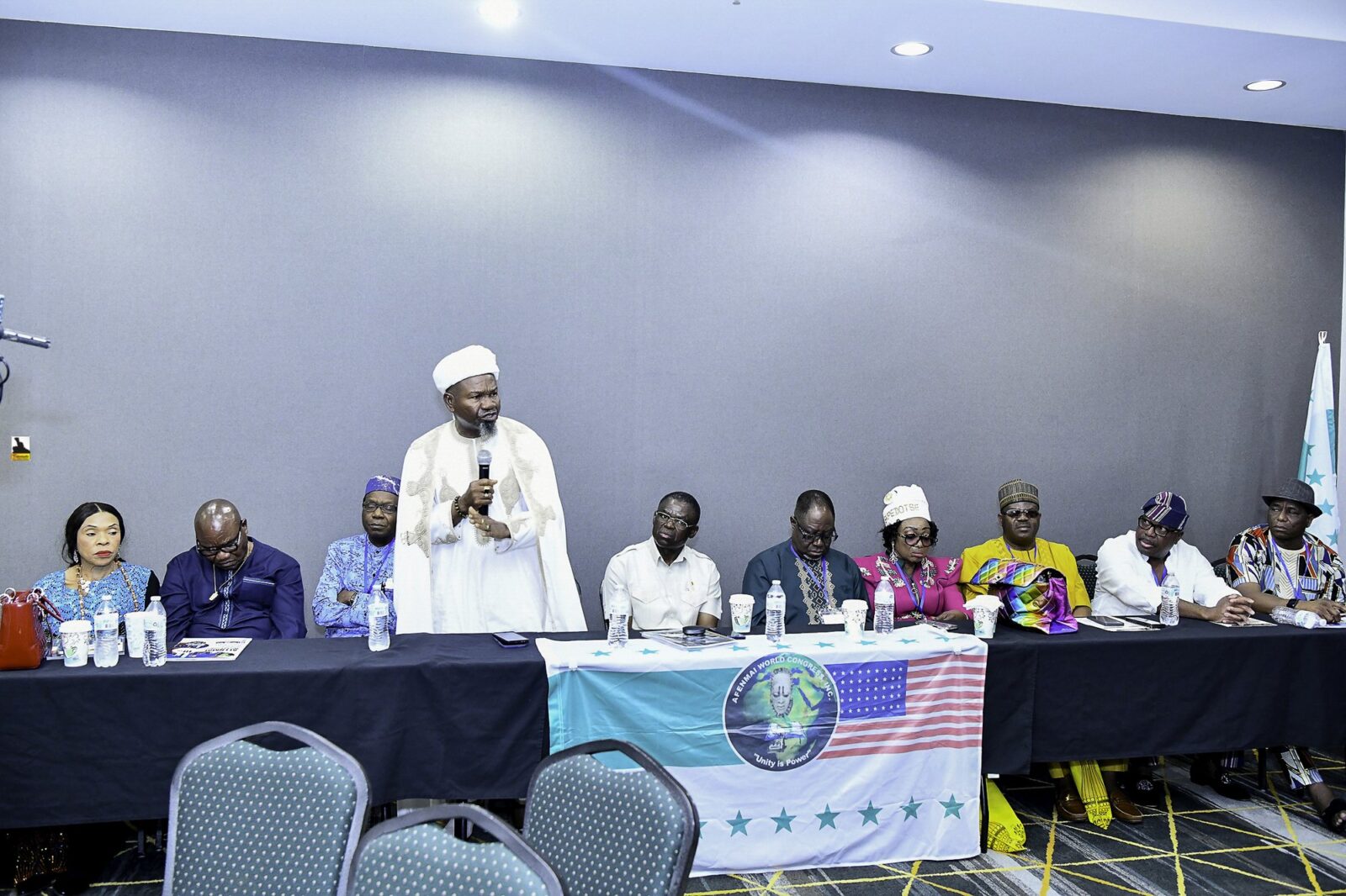 Man in embroidered white clothing addressing attendees at the front table