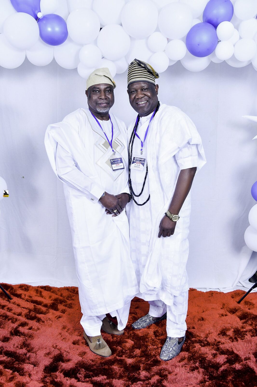 Two men in white clothing posing beside the balloon backdrop