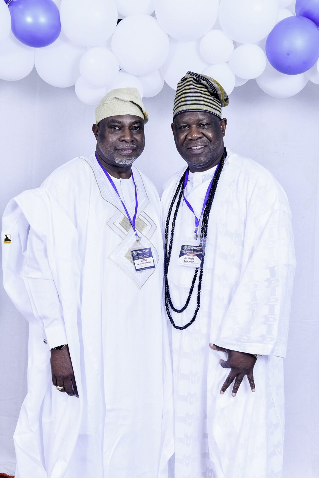 Two men in white clothing posing beside the balloon backdrop