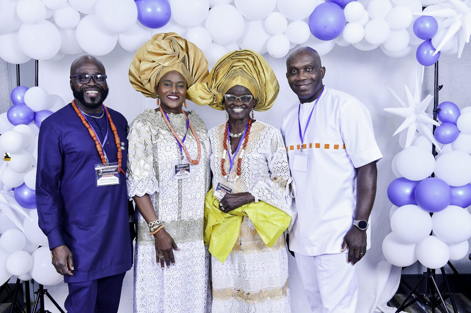 Four smiling attendees in blue and white, with gold headwraps, beneath balloons