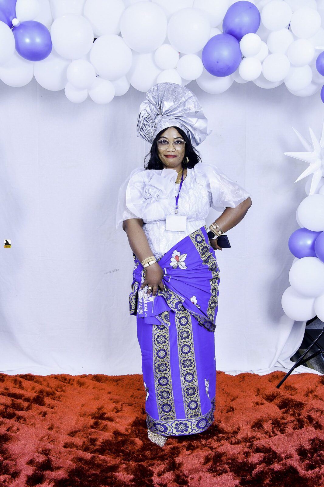 Woman in a white blouse, blue wrapper and silver headwrap posing by the balloons