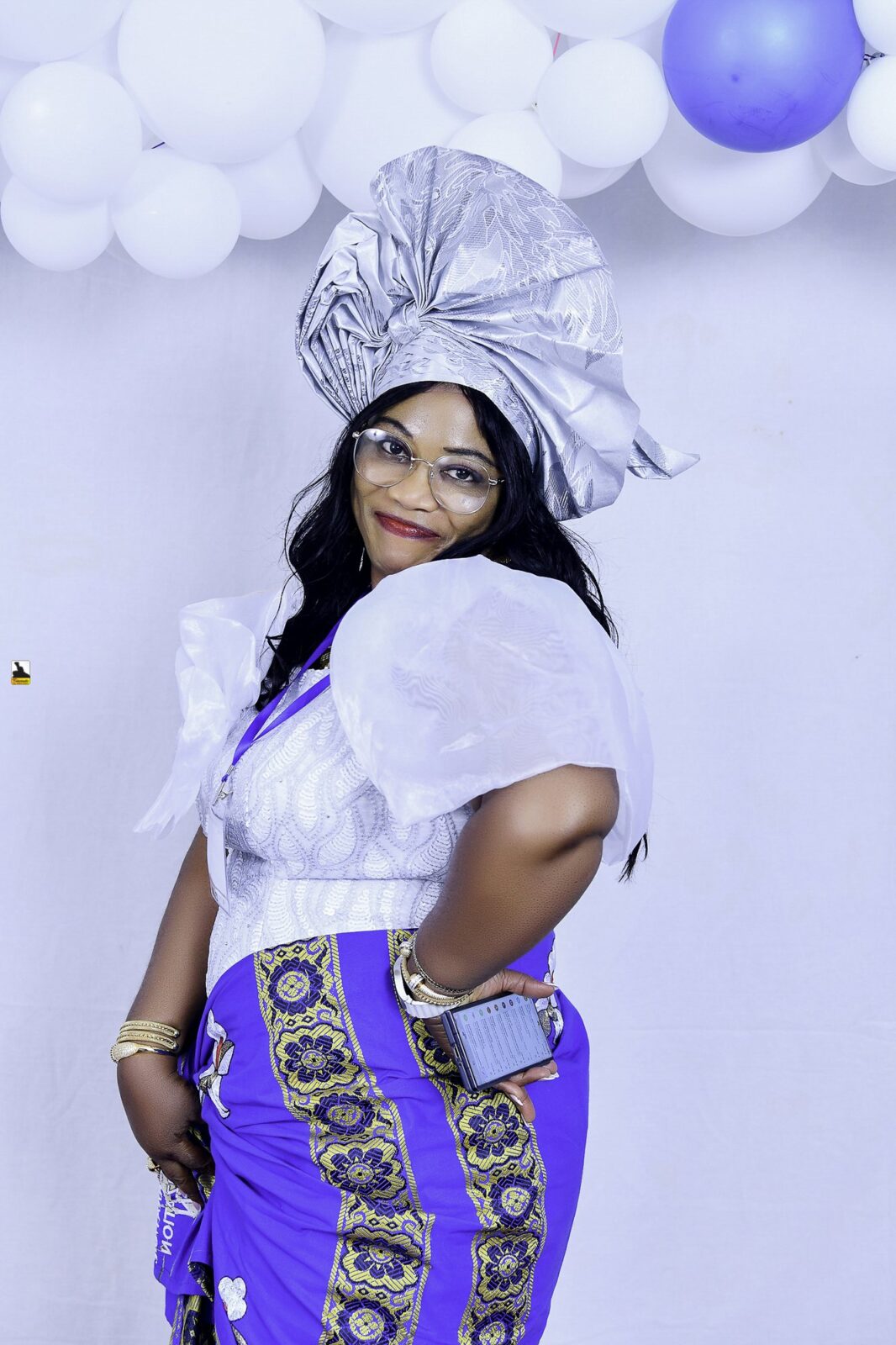 Woman in a white blouse, blue wrapper and silver headwrap posing by the balloons