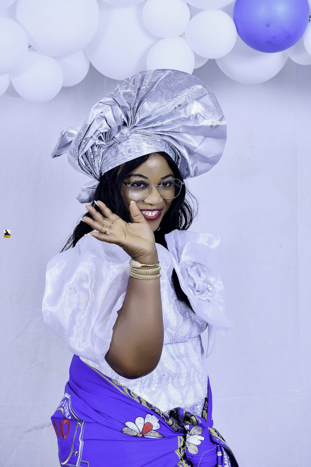 Woman in a white blouse, blue wrapper and silver headwrap posing by the balloons