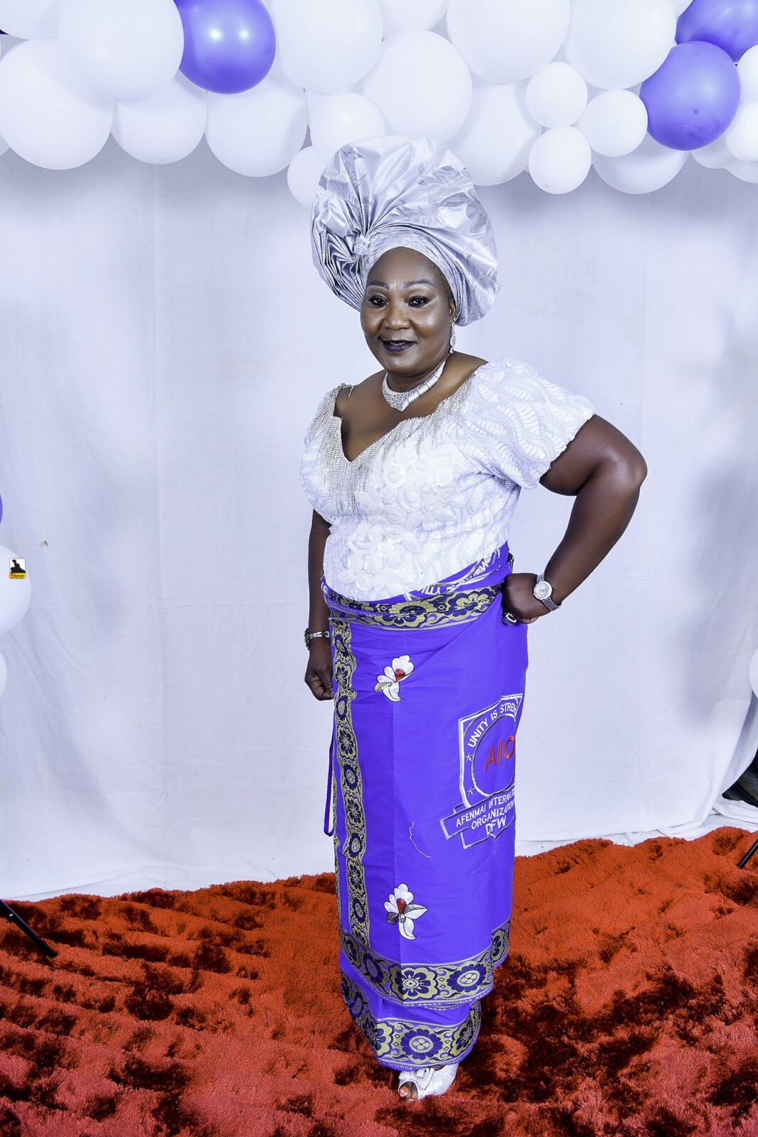 Woman in a white blouse, blue wrapper and silver headwrap posing by the balloons