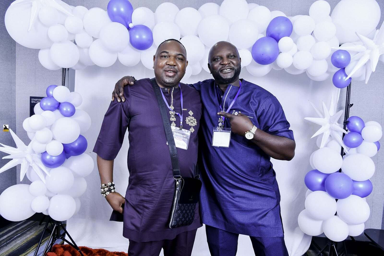 Two men in blue outfits smiling together beneath the balloons