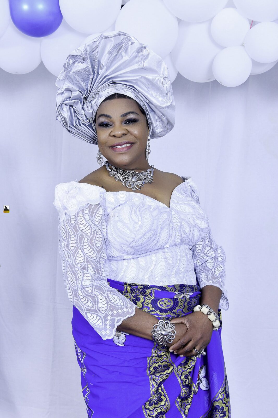 Woman in white lace, a blue wrapper and a silver headwrap by the balloons