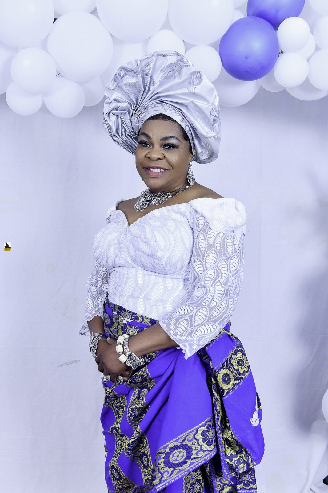 Woman in white lace, a blue wrapper and a silver headwrap by the balloons