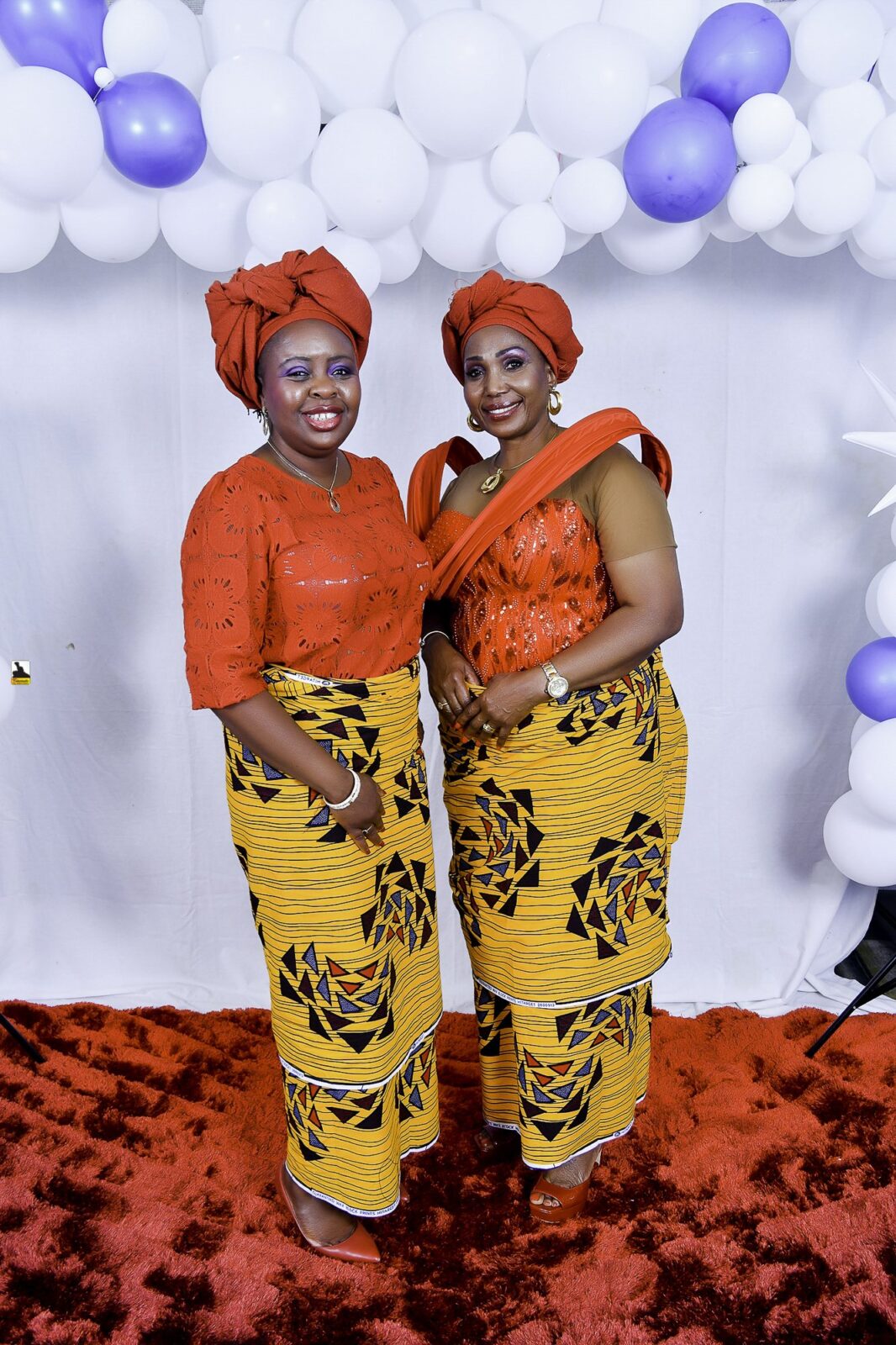 Two women in orange tops, yellow patterned wrappers and orange headwraps