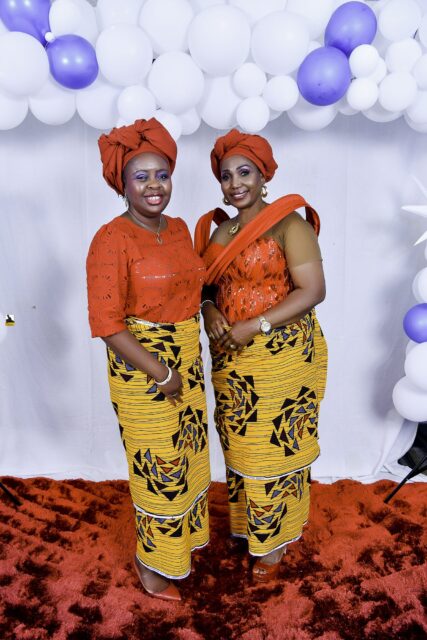 Two women in orange tops, yellow patterned wrappers and orange headwraps