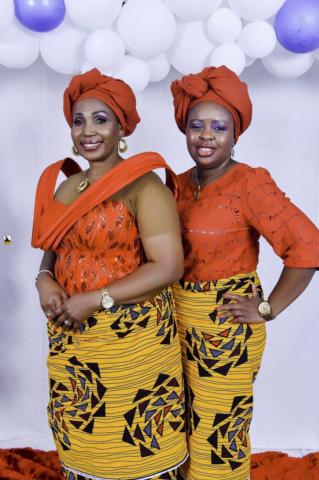 Two women in orange tops, yellow patterned wrappers and orange headwraps