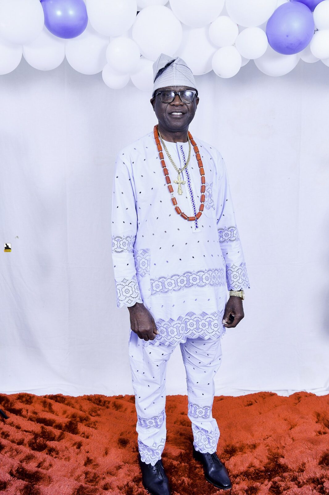 Man in white patterned clothing and bead necklaces by the balloons