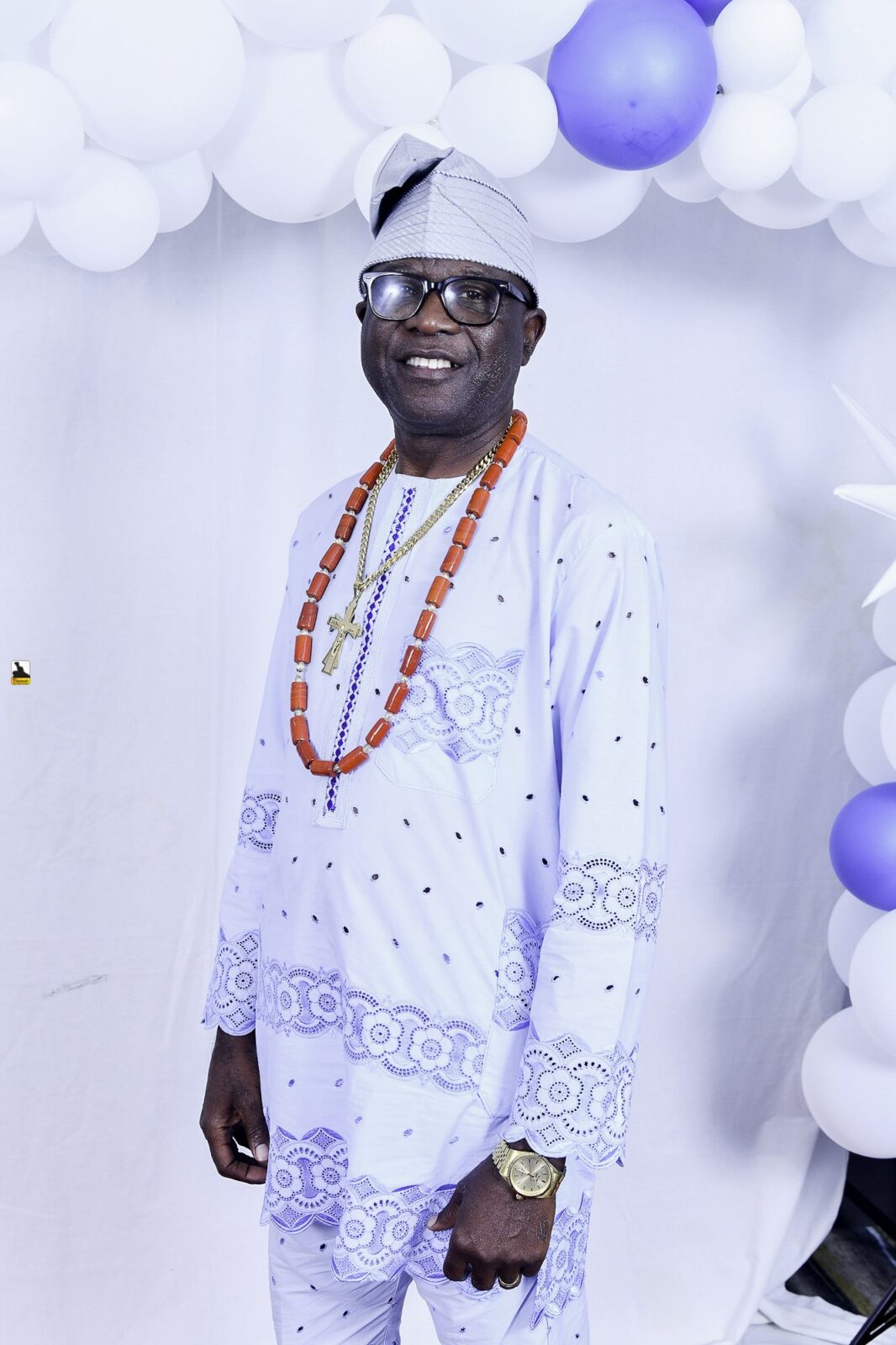 Man in white patterned clothing and bead necklaces by the balloons