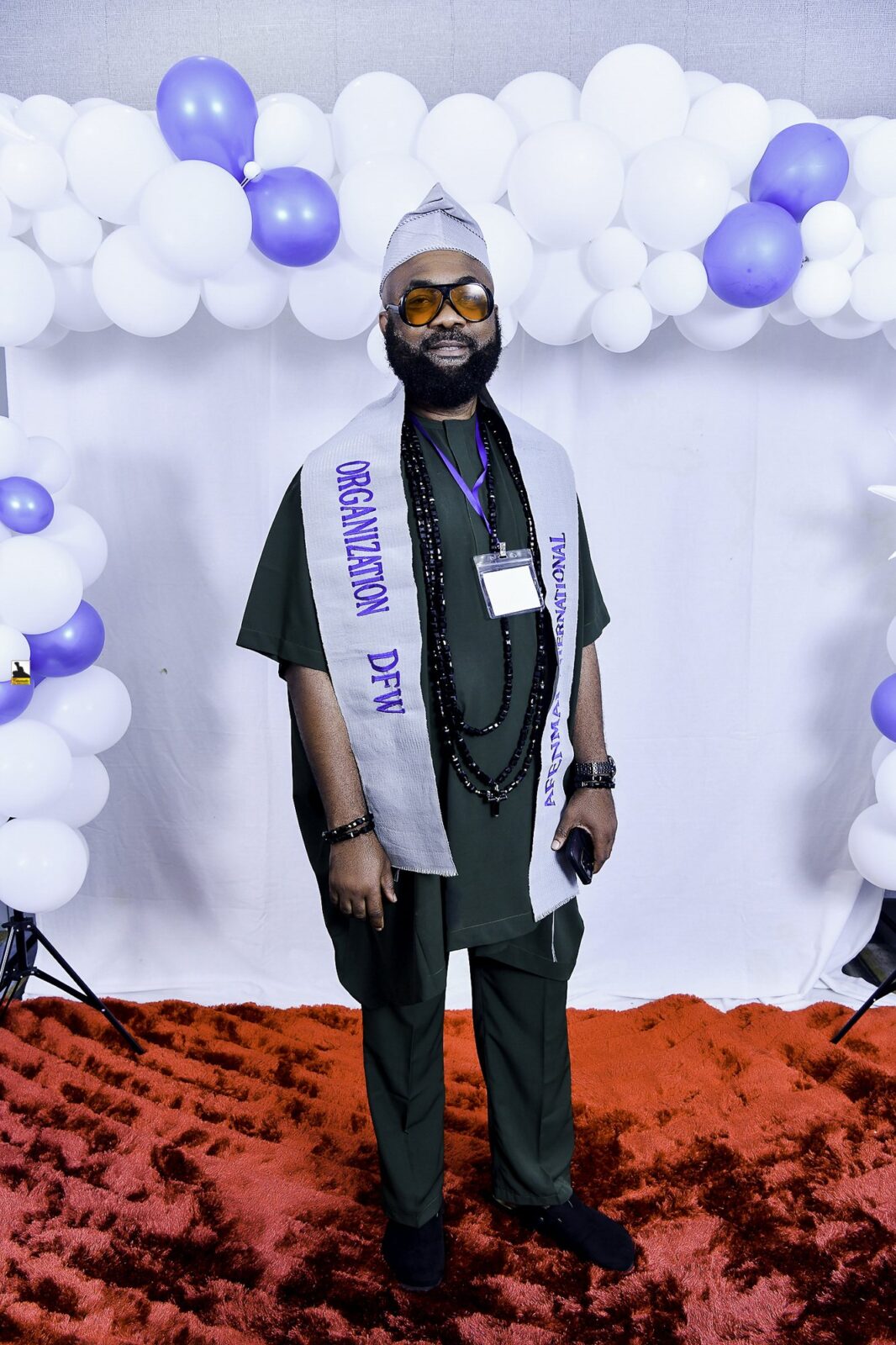 Man in dark clothing, glasses and a lettered sash by the balloons