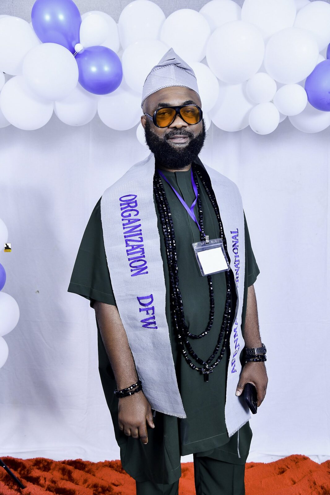 Man in dark clothing, glasses and a lettered sash by the balloons