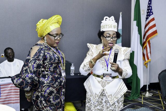 Two women standing together, one holding a microphone beside the flags