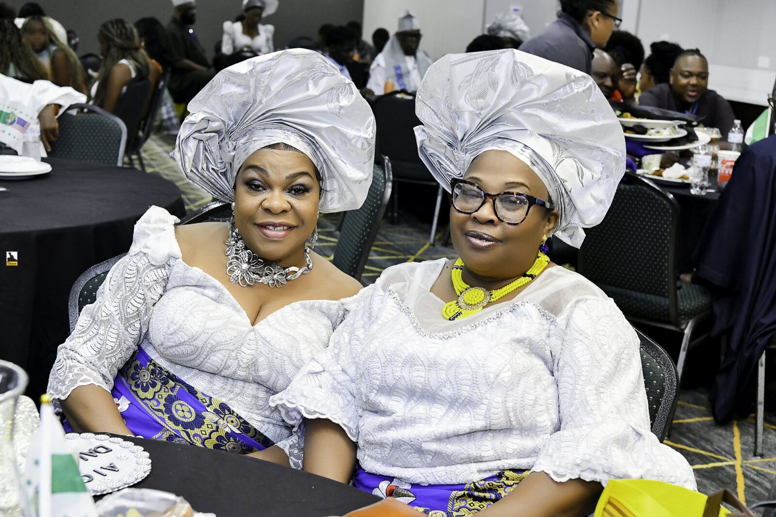Two smiling women in white lace and silver headwraps seated together