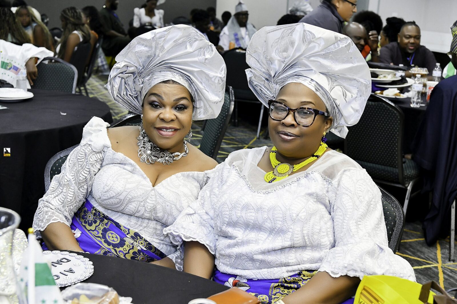 Two smiling women in white lace and silver headwraps seated together