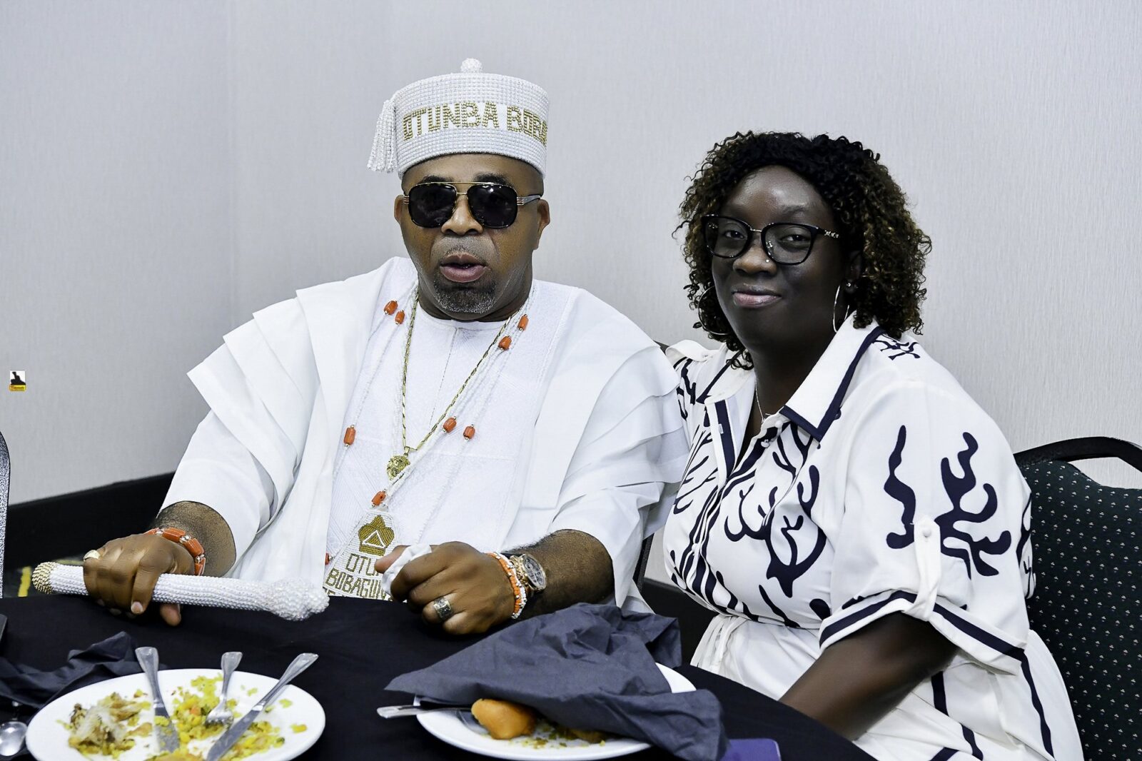 Man in white with sunglasses and woman in a patterned top at a table