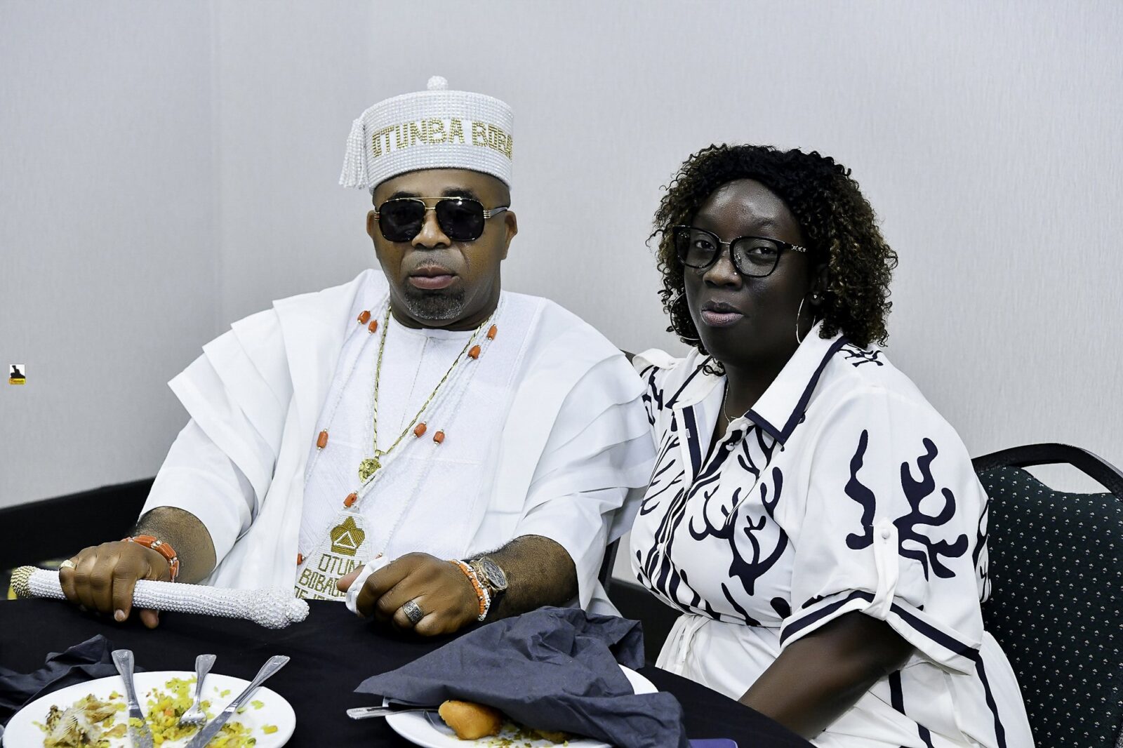 Man in white with sunglasses and woman in a patterned top at a table