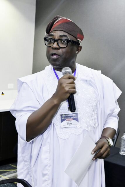 Man in pale clothing and a red cap speaking into a microphone